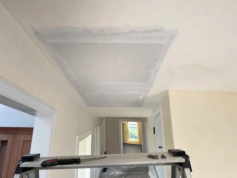 A hallway ceiling where a water-stained section of drywall was cut out and a new piece screwed in, the seams taped and first-coated