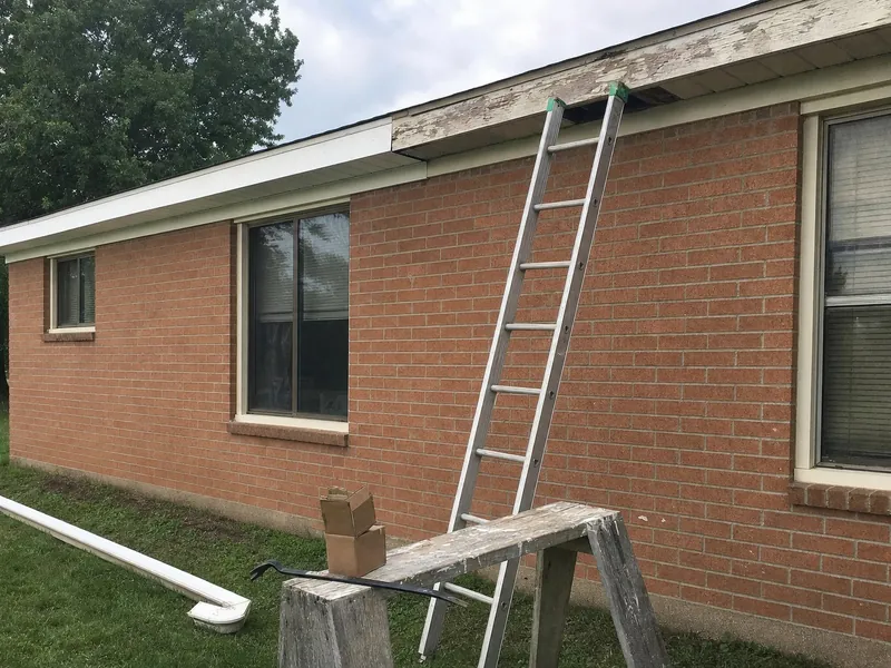 The eave of a brick ranch house: new white fascia and vented soffit next to old weathered wood still to be replaced, a ladder against the wall