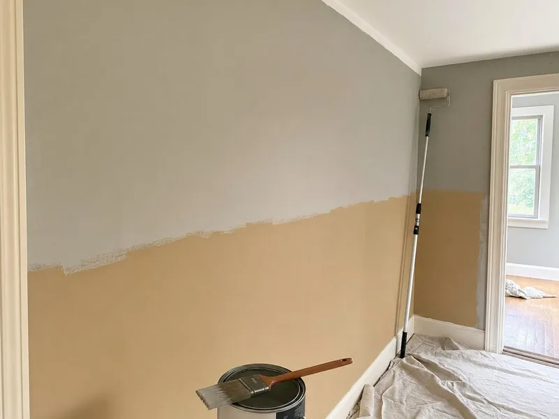 A hallway halfway through a new coat of paint: the top of the wall in a new soft grey, the bottom still tan, a brush resting on an open paint can