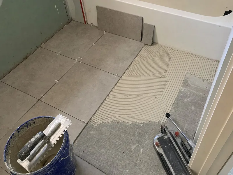 A bathroom floor half set in grey porcelain tile with spacers, the rest bare backer board with thinset combed in lines